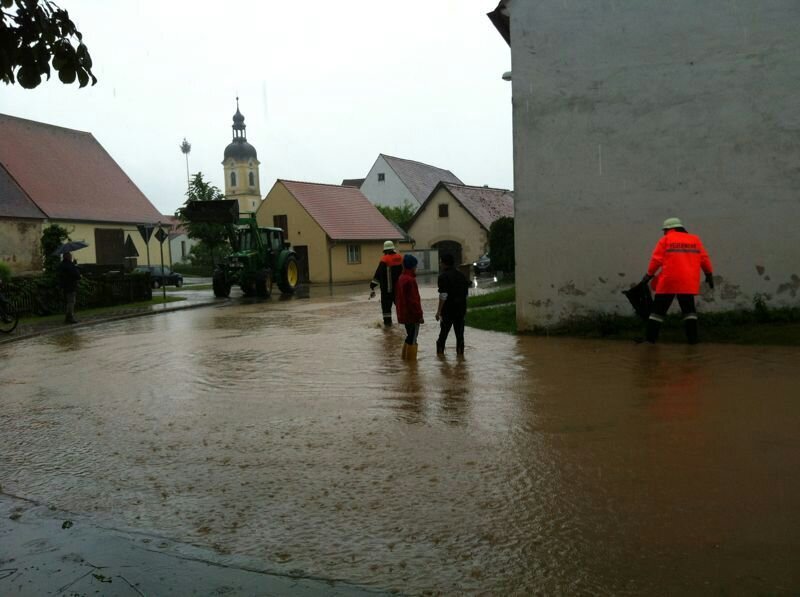 Überflutung Stopfenheim Eine überflutete Straße in einer Gemeinde. Vier Menschen watten durch das Wasser, das ihnen bis zum Schienbein reicht.