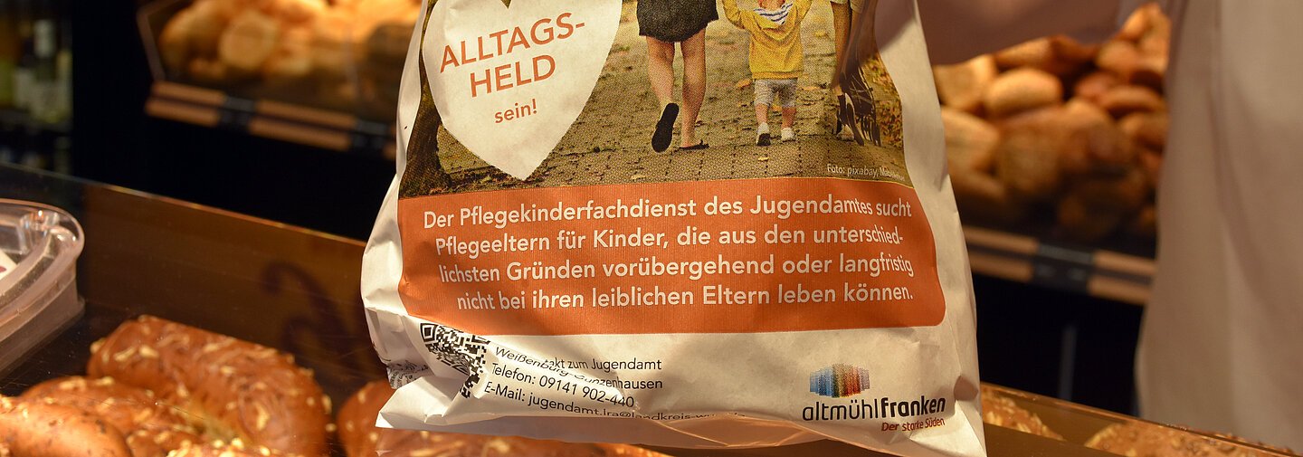 Aktionsbäckertüte zum Thema "Pflegeeltern werden" wird über die Verkaufstheke einer Bäckerei gereicht.