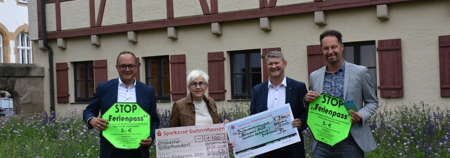 Drei Männer und eine Frau präsentieren mit Schildern und einem Spendenscheck den Start der Aktion Ferienpass 2025.