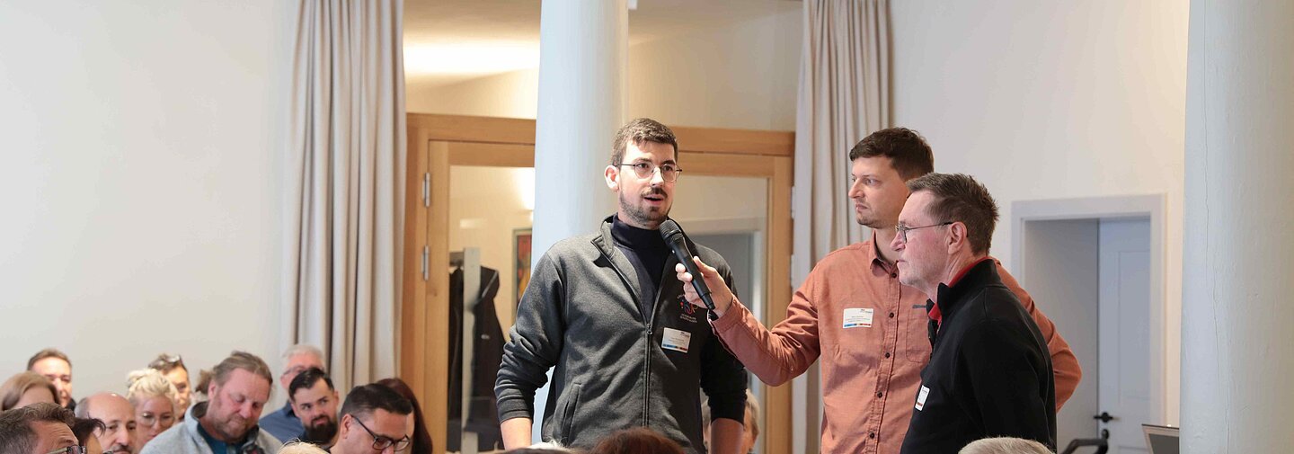 Ein Mann wird von einem anderen Mann in einem Raum voller Menschen interviewt.