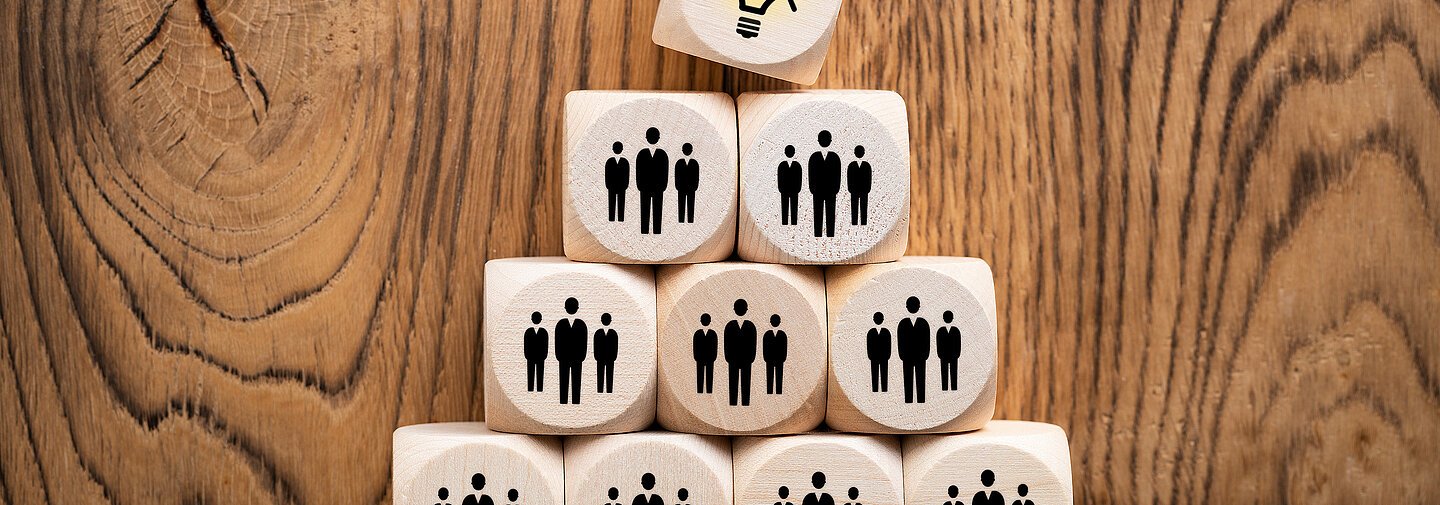 Idee durch Teamwork Würfel mit Menschen als Augen liegen pyramidenförmig auf einem Holzhintergrund. Die Spitze ist ein Würfel, der eine Glühbirne als Auge hat.