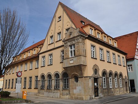 Historisches Gebäude mit gelber Fassade, roten Dachziegeln und Erker an Straßenecke bei bewölktem Himmel.