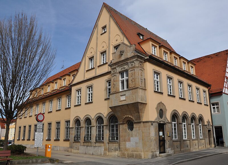 Historisches Gebäude mit gelber Fassade, roten Dachziegeln und Erker an Straßenecke bei bewölktem Himmel.