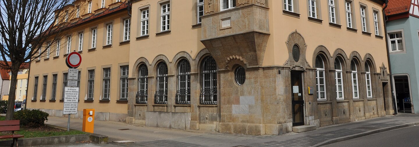 Historisches Gebäude mit gelber Fassade, roten Dachziegeln und Erker an Straßenecke bei bewölktem Himmel.