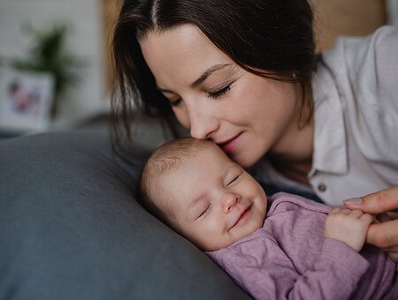 A happy young mother kissing her newborn baby girl, lying on sofa indoors at home.